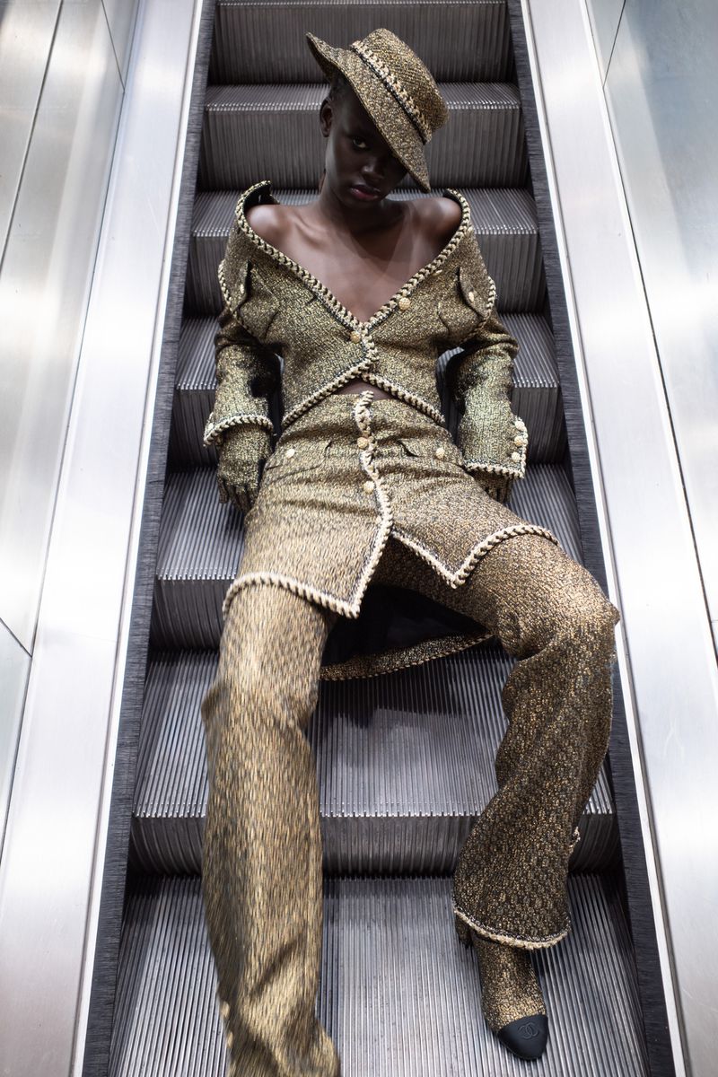 Model seated on metal escalator wearing a metallic tweed suit and matching hat for Heroine Magazine.
