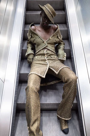 Model seated on metal escalator wearing a metallic tweed suit and matching hat for Heroine Magazine.