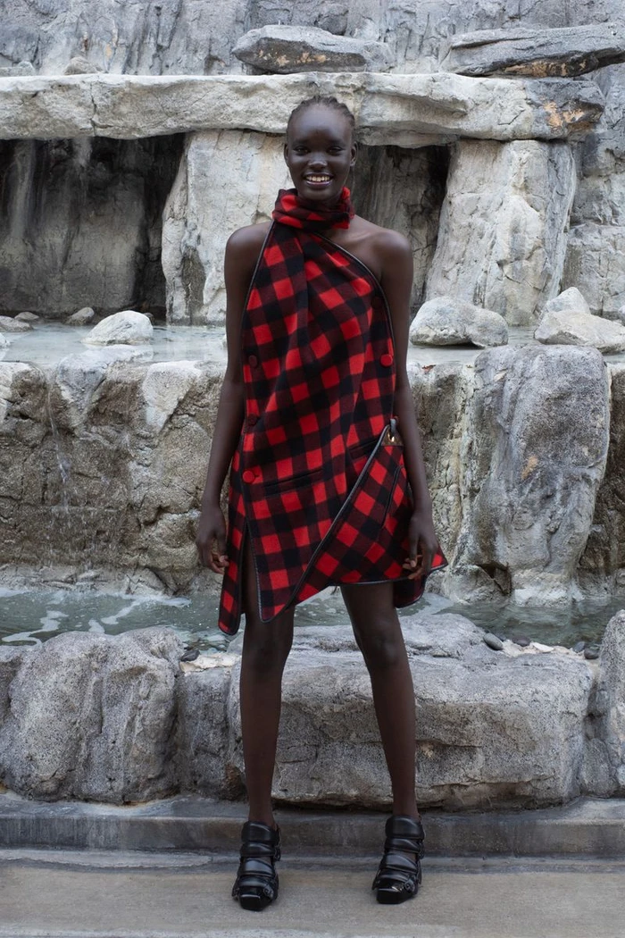 Model in red and black plaid dress standing against a textured rock formation and water feature