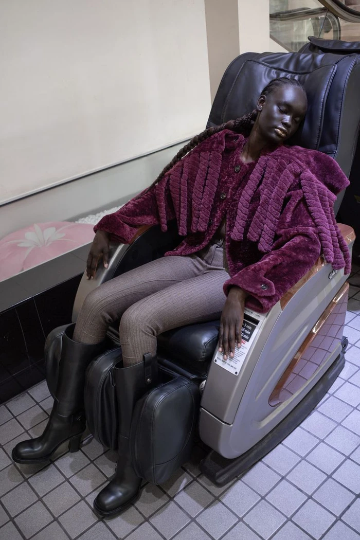 Model wearing plum textured jacket and tall black boots resting in a massage chair for Heroine Magazine