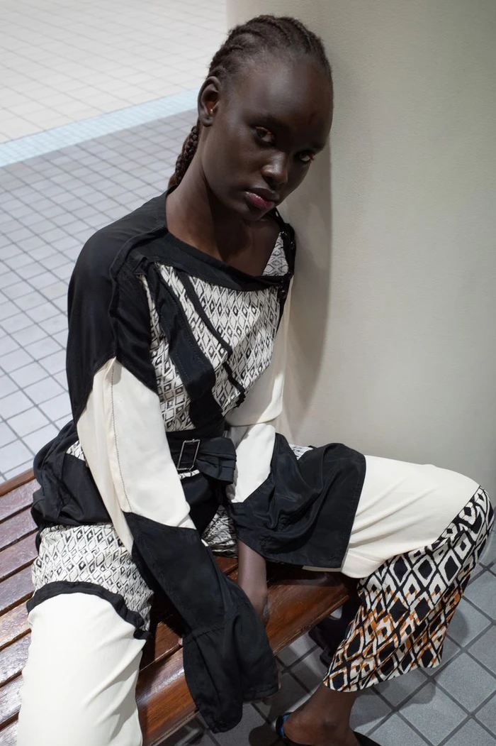 Model Alek Maduk seated on a wooden bench wearing patterned monochrome apparel for Heroine Magazine