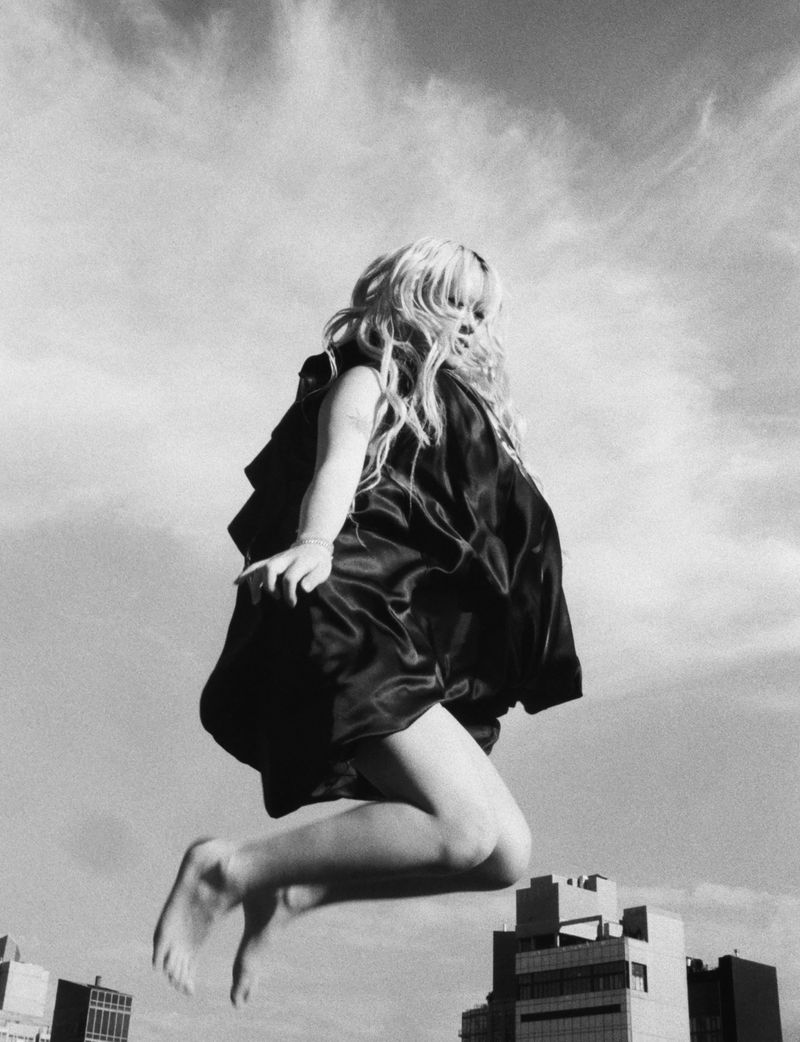 Black and white photo of a young woman jumping in the air wearing a dress against a sky backdrop with buildings below.