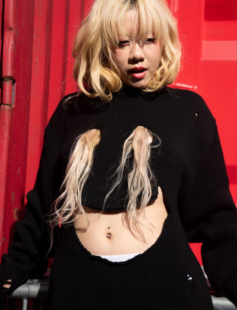 Blonde woman in a black, distressed sweater with cut-outs posing against a red metal wall, showcasing street-style fashion.