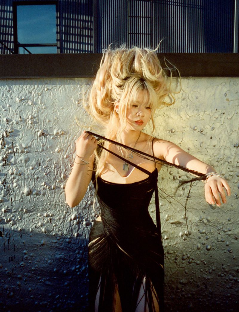 Young adult female model in a black dress posing outside in front of an textured wall with a modern building with slatted windows.