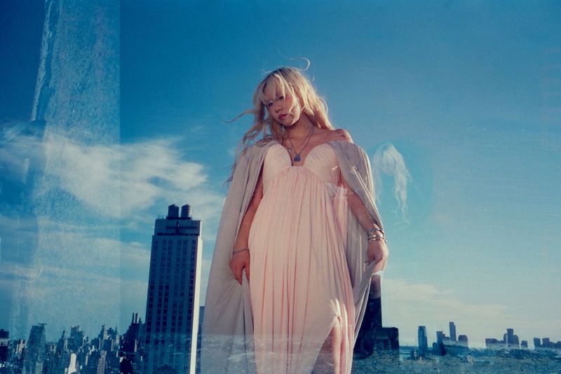Blonde woman in a light pink dress and cape standing on a rooftop with a cityscape and blue sky background in a fashion photoshoot.