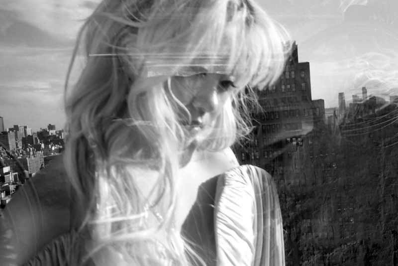 Black and white portrait of a woman with blond hair in front of a city skyline, blending the figure with the urban background.