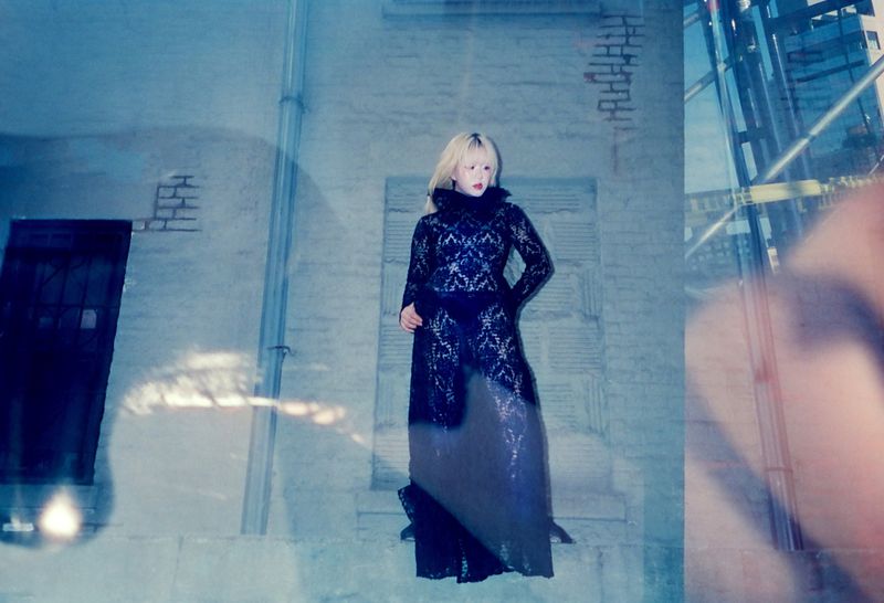Fashion photography of a woman with blonde hair wearing a long black lace dress against a brick wall backdrop, captured in an urban environment.