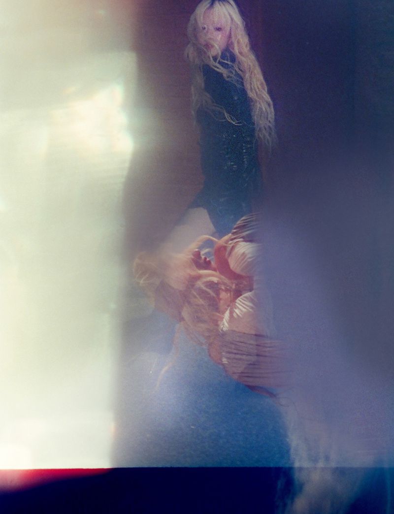 Blonde adult female poses in a blurred, stylized fashion shot with cool tones, creating an ethereal and artistic photographic composition.