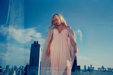 Blonde woman in a pale pink dress and draped cape against a city skyline with multiple exposure reflections