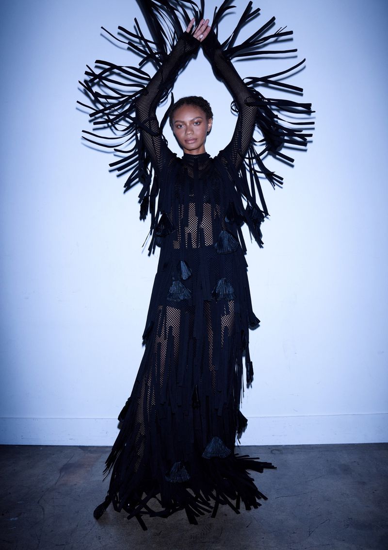 Woman in a black fringed dress raises her arms in a fashion pose in front of a light blue wall with a dark gray floor.