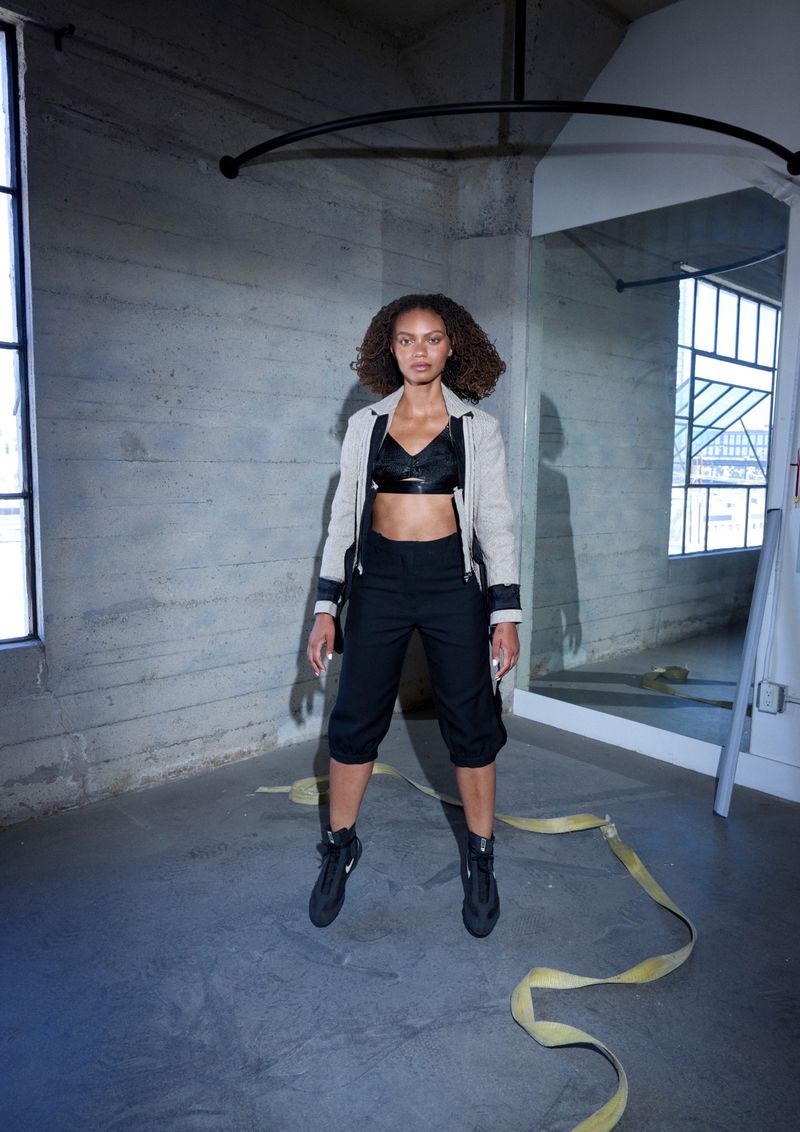 Fashion portrait of a woman in a blazer and black crop top. She stands in an empty room with concrete walls and a yellow ribbon.