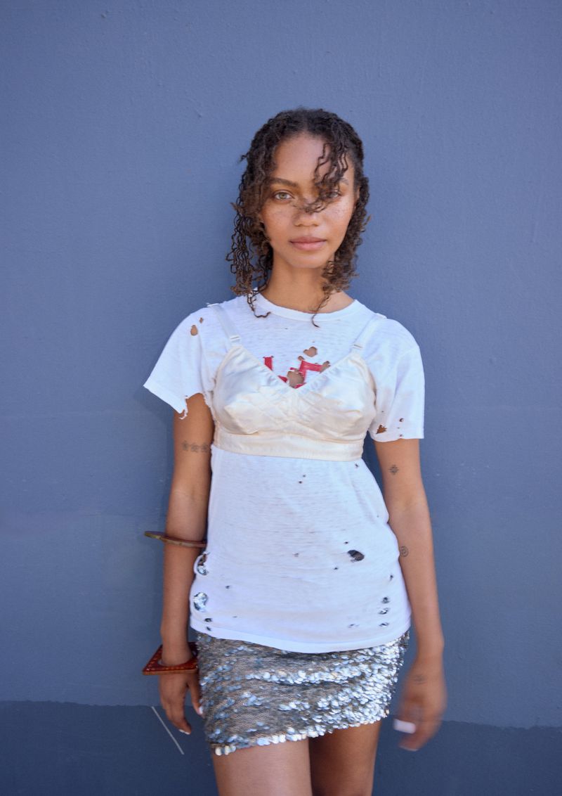 Young adult woman posing in a distressed white t-shirt and sequined mini-skirt against a muted blue wall in an eye-level medium shot.