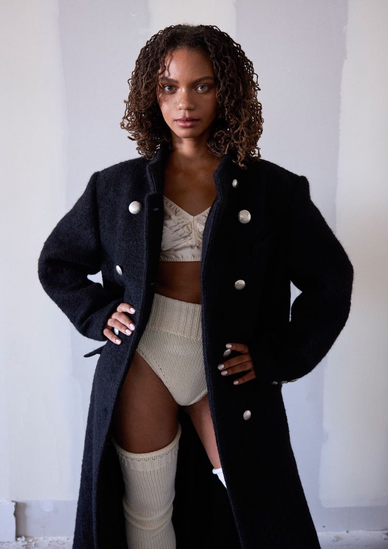 Portrait of a woman in an unbuttoned black coat, knit underwear, and thigh-high socks posing in a photo studio with a neutral backdrop.