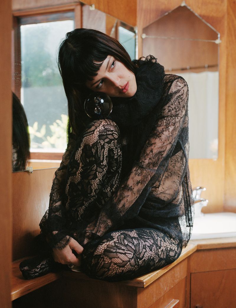 Young woman with black hair in lace top sits in front of vanity with orb, bathed in soft, natural light in an indoor setting.