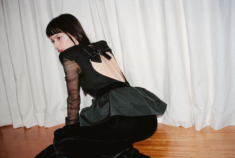 Brunette woman in a black gown with long sleeves and an open back, posing on a wood floor in front of a white curtain.