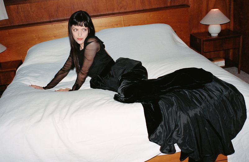 Woman in a long black dress poses on a bed in a bedroom. The woman has black hair and is lying on a white bedspread.