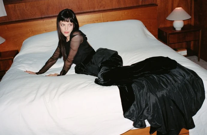 Woman in a long black dress poses on a bed in a bedroom. The woman has black hair and is lying on a white bedspread.