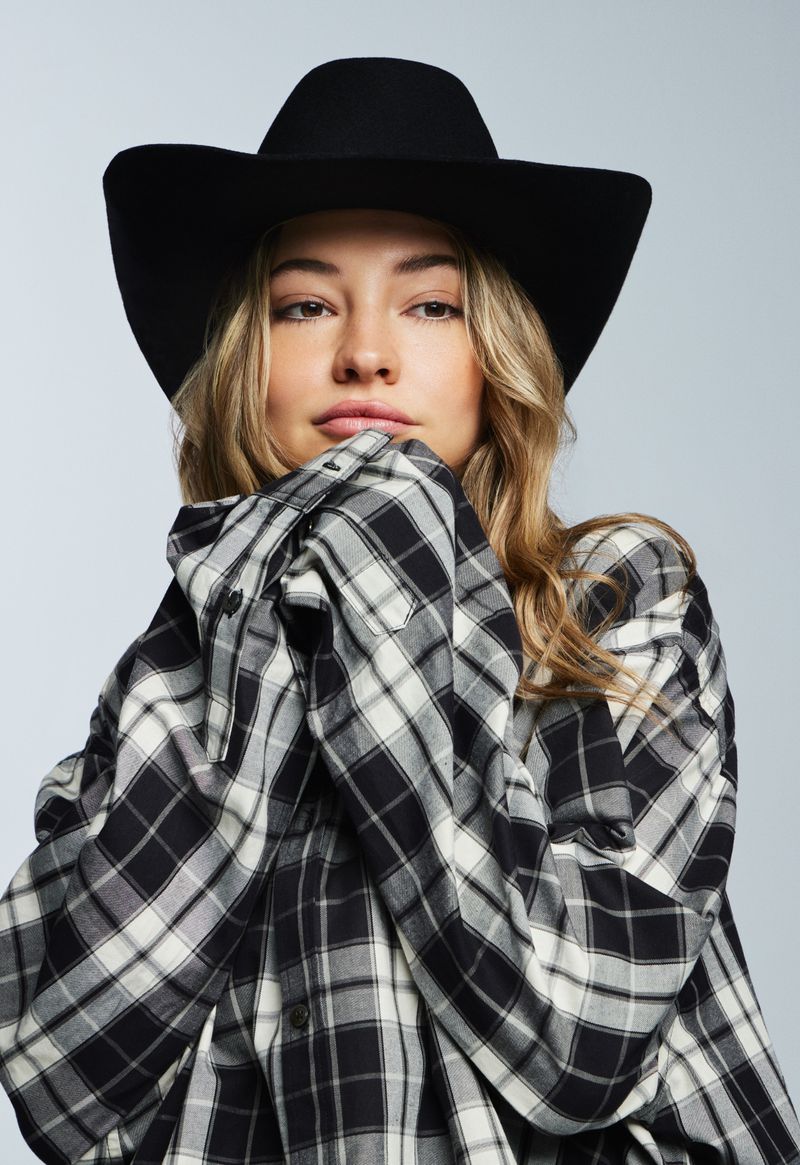 Portrait of a young woman in a black cowboy hat and an oversized black and white plaid shirt posing in front of a gray background.