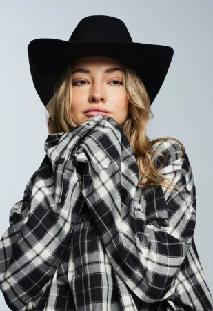 Portrait of a young woman in a black cowboy hat and an oversized black and white plaid shirt posing in front of a gray background.