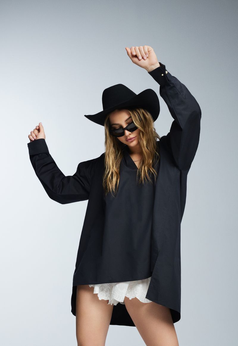 Fashionable woman in black cowboy hat, oversized shirt, and lace shorts dancing against a gradient gray studio backdrop, arms raised.