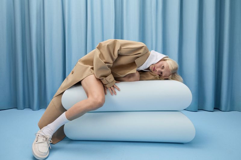 Fashion shot of a blonde adult woman in a tan coat, white hoodie and socks, and cream shoes, lying on a stacked light-blue cylinder set in a photo studio.