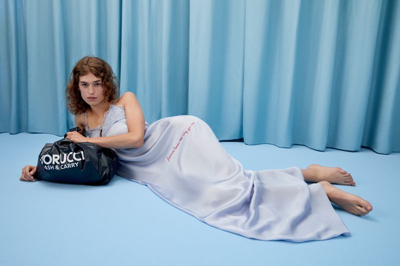 A woman lounges in a light blue gown in front of matching curtains, beside a black Fiorucci shopping bag, indoors with soft lighting.