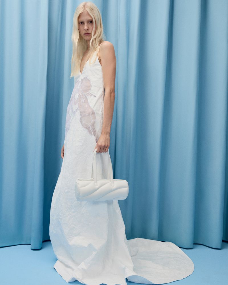Woman in a white dress and blonde hair poses with a white purse against a light blue curtain in a studio, fashion editorial shot.