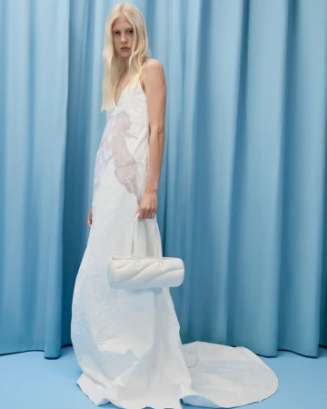Woman in a white dress and blonde hair poses with a white purse against a light blue curtain in a studio, fashion editorial shot.