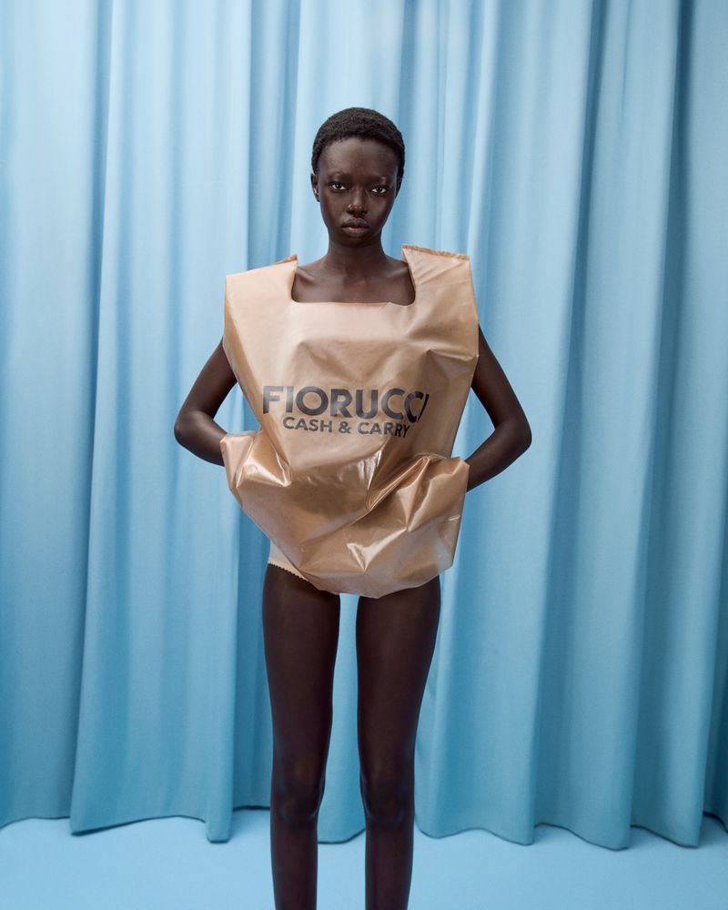 A slender, dark-skinned woman in a tan shopping bag dress stands against a light-blue curtain backdrop in a fashion-forward portrait.