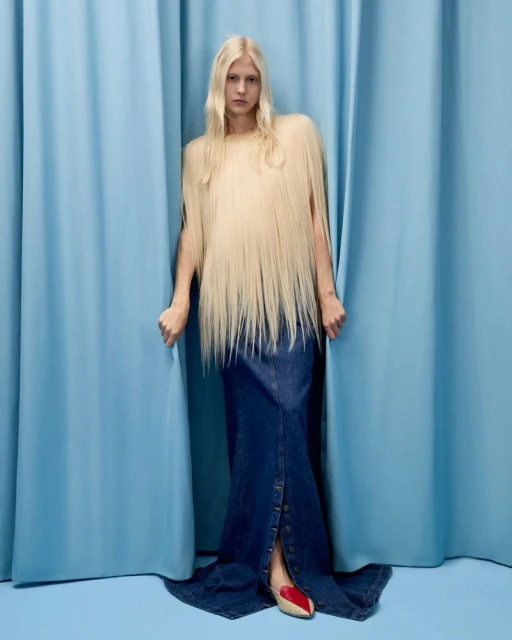Woman with long blonde hair wears a long denim skirt and stands between blue curtains in a fashion studio portrait. 
