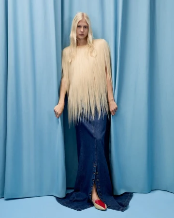 Woman with long blonde hair wears a long denim skirt and stands between blue curtains in a fashion studio portrait. 