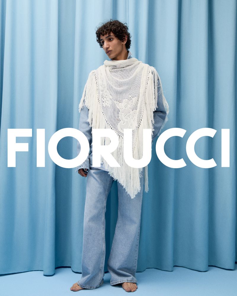 Full-length shot of a person wearing light-wash denim and a white shawl against a light blue draped fabric backdrop, fashion shoot.