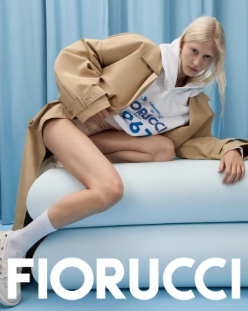 Fashion editorial shot of a blonde adult female in a tan trench coat, hoodie, and white sneakers posing on blue cylinders.