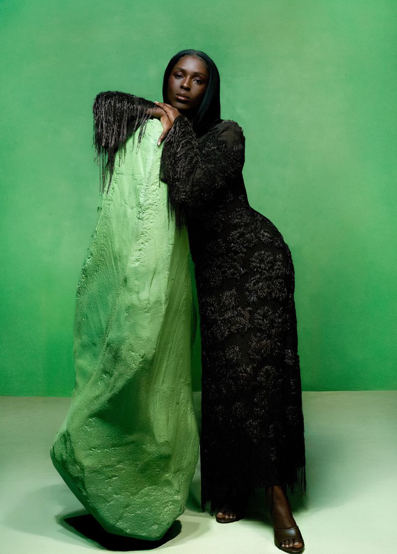 Elegant Black woman in long black dress leaning against abstract light-green stone sculpture in studio with bright green background.