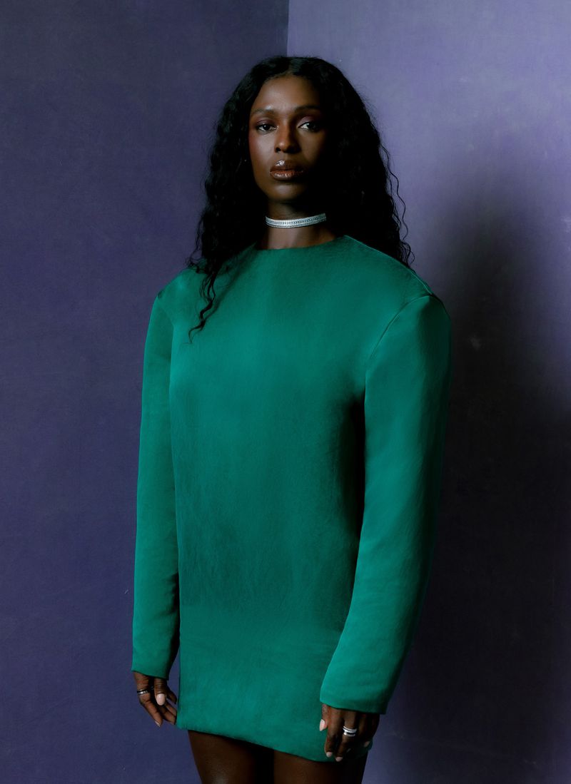 Portrait of an adult woman with dark hair and skin wearing a green dress against a textured purple wall inside a photo studio.