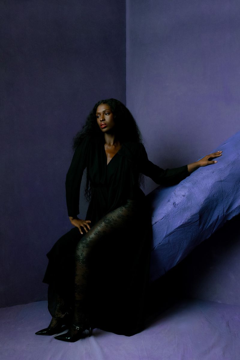 Elegant woman in long black dress poses against a textured blue wall in a studio setting. Sophisticated fashion shot with dramatic lighting.