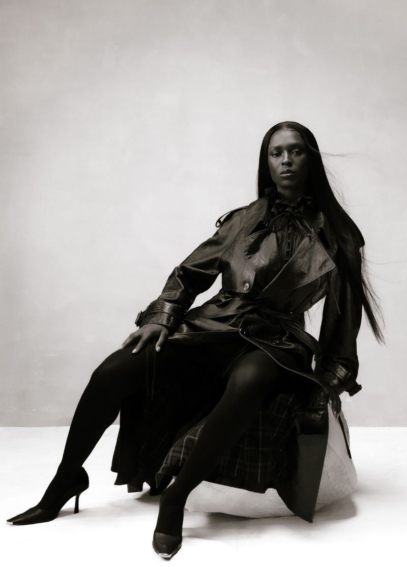 A dark-haired woman sits in a leather trench coat, striking a pose in a monochrome fashion shot against a neutral backdrop, exuding elegance.