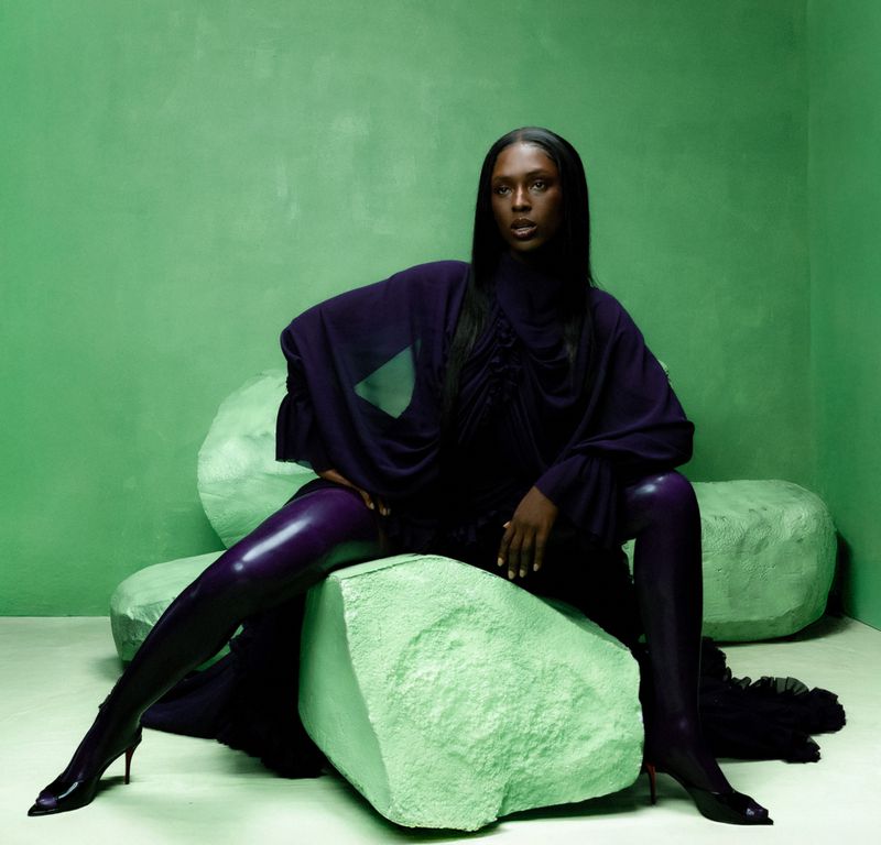 A striking studio portrait of a Black female model with dark hair wearing a dark purple dress and matching shiny leggings, posing on light green rocks.