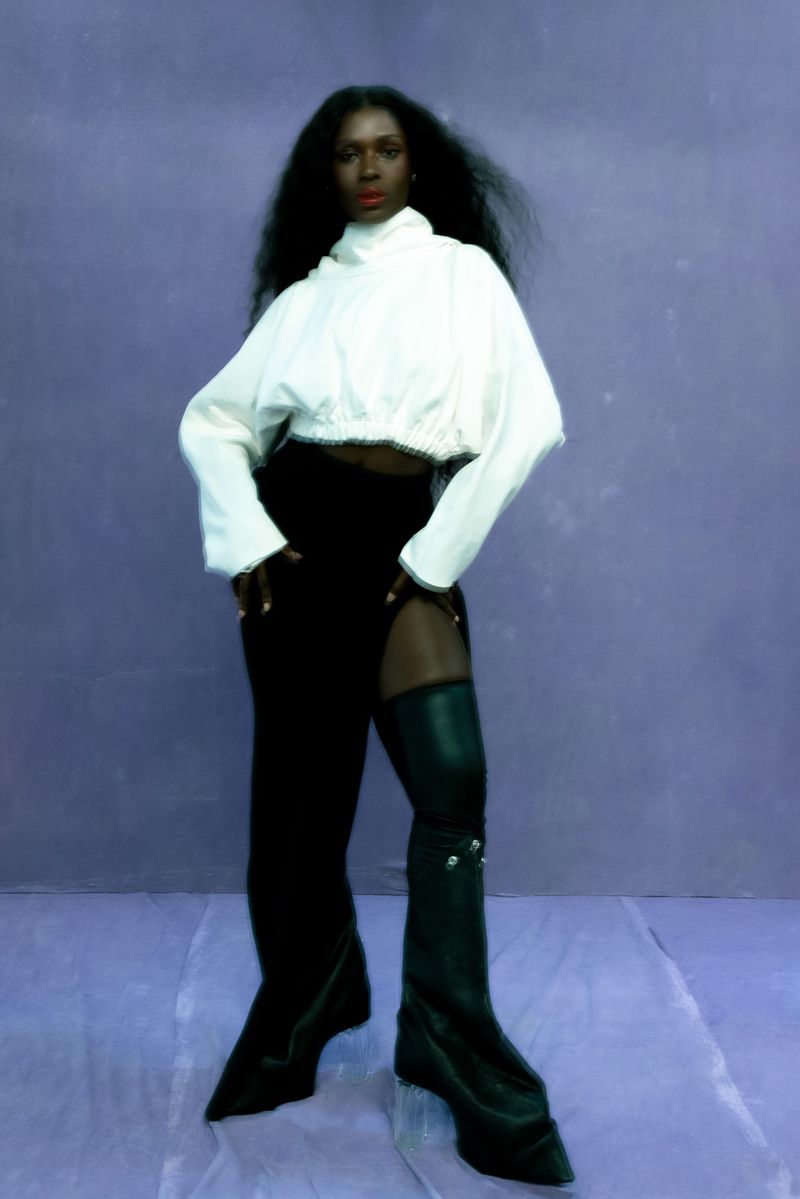 Elegant Black woman in a high-fashion pose against a purple background wearing a white top, black skirt and leather boots.