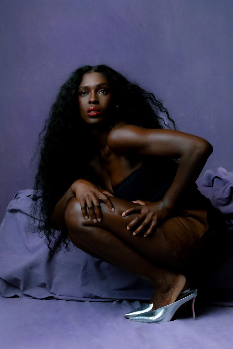 Elegant African American woman with long dark curly hair posing in a crouched position on a violet draped surface, wearing heeled shoes.