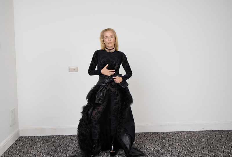Gillian Anderson in long-sleeved black gown with feathered trim, standing in interior space with patterned carpet, full body shot.