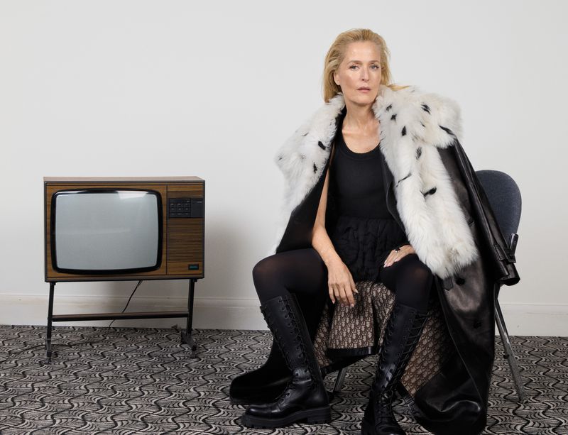 Stylish mature woman in black dress, lace-up boots, and fur-trimmed coat sits on a chair beside a vintage television in a studio.