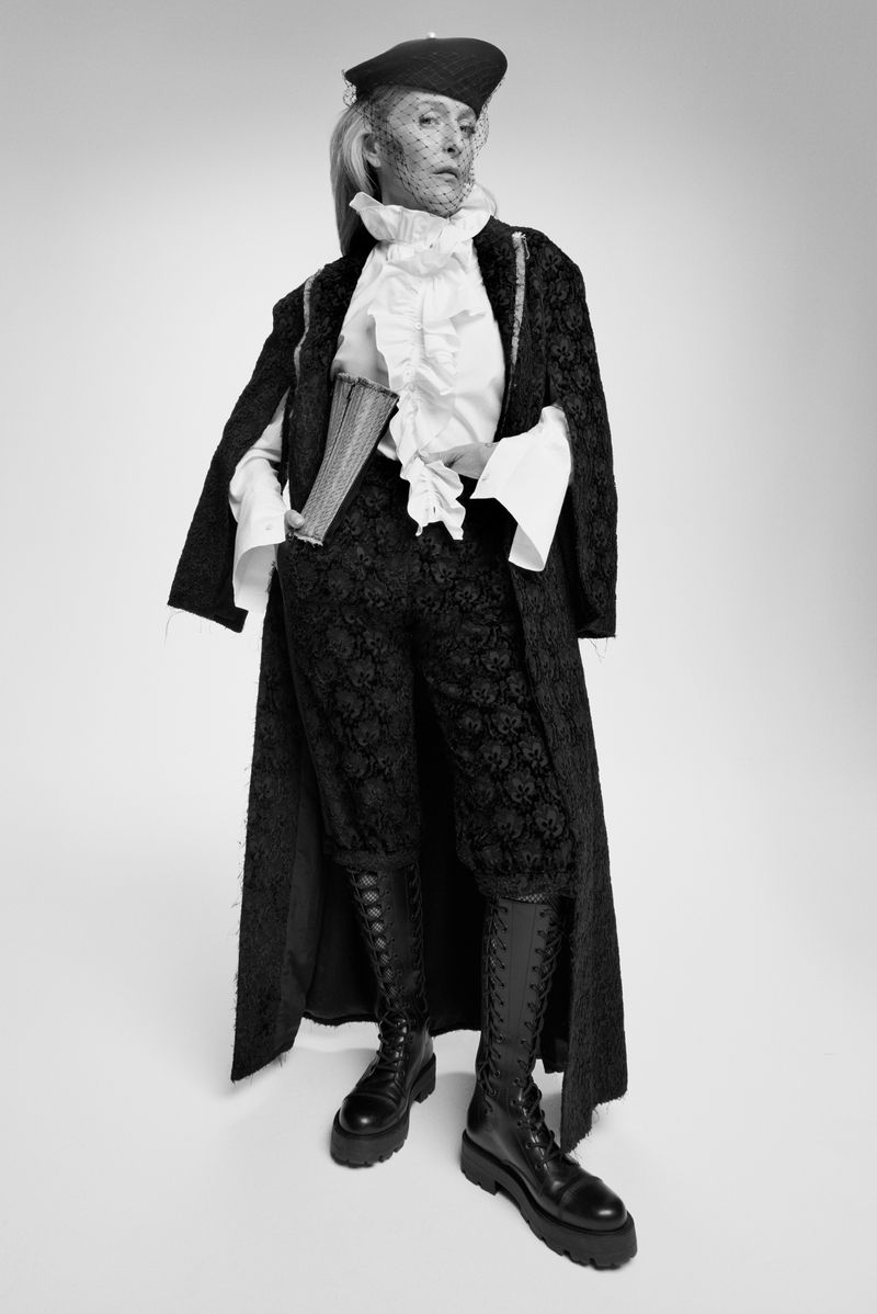 Full-length shot of a woman in a vintage-style black patterned suit, frilled white shirt, black boots, and a black beret with net veil on a white background.