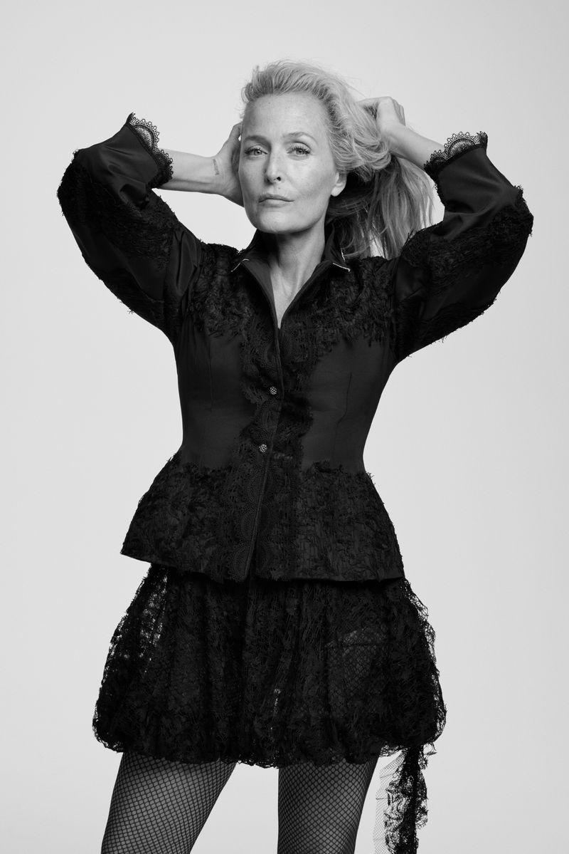Mature woman in black lace top and skirt poses indoors with hands in her hair in black and white for a fashion editorial.