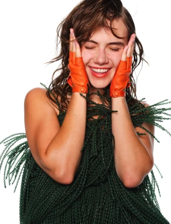 Emilia Jones laughing with eyes closed wearing orange leather fingerless gloves and a textured green garment against a white background.