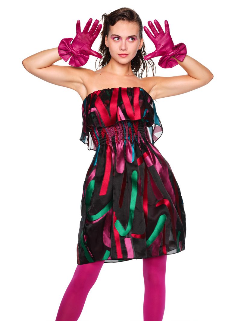 A young adult female fashion model poses in a colorful strapless dress and magenta gloves and leggings against a white backdrop.