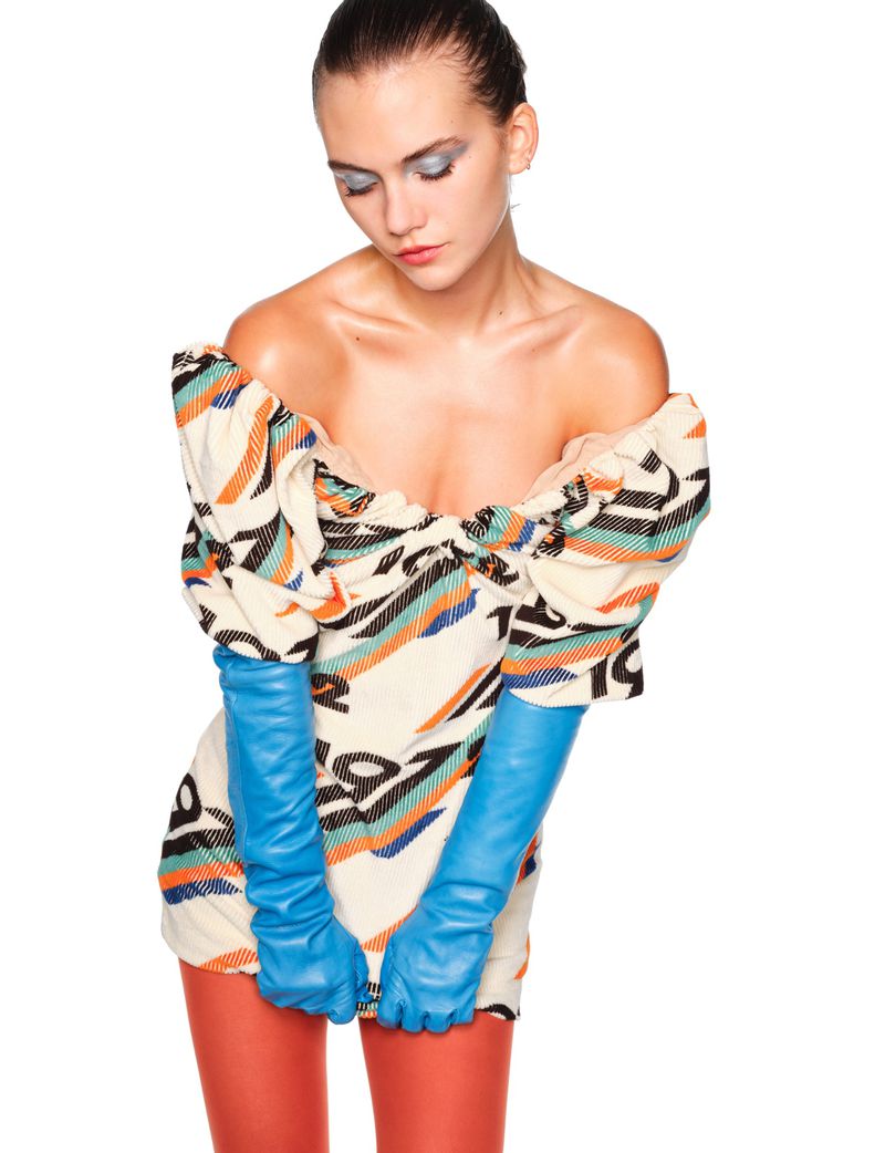 Fashion shot of a woman in a patterned dress, long blue gloves, and red tights against a bright white background. Makeup is clean and modern.