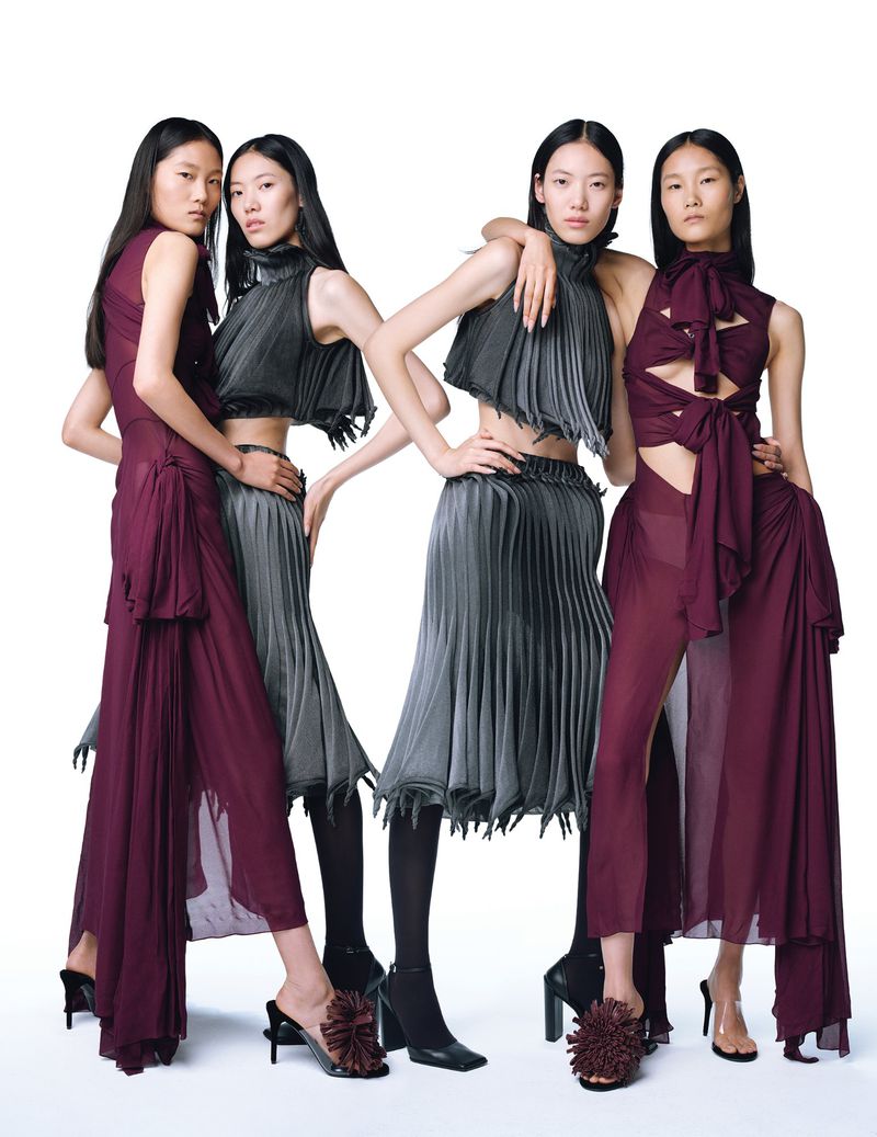 Four Asian fashion models pose in a studio wearing maroon and grey pleated two-piece outfits, high neck tops, skirts, and high heeled shoes.