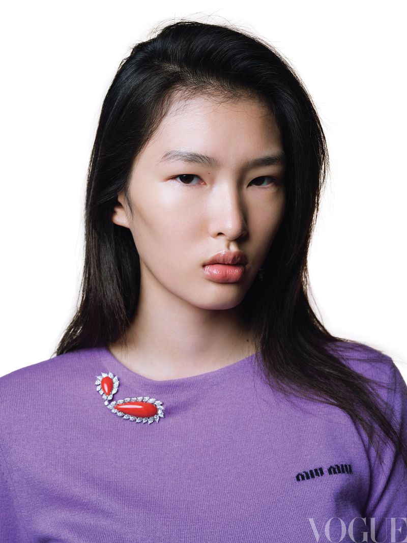 Close-up studio shot of a young Asian woman with dark hair and fair skin wearing a lilac sweater with a decorative brooch.