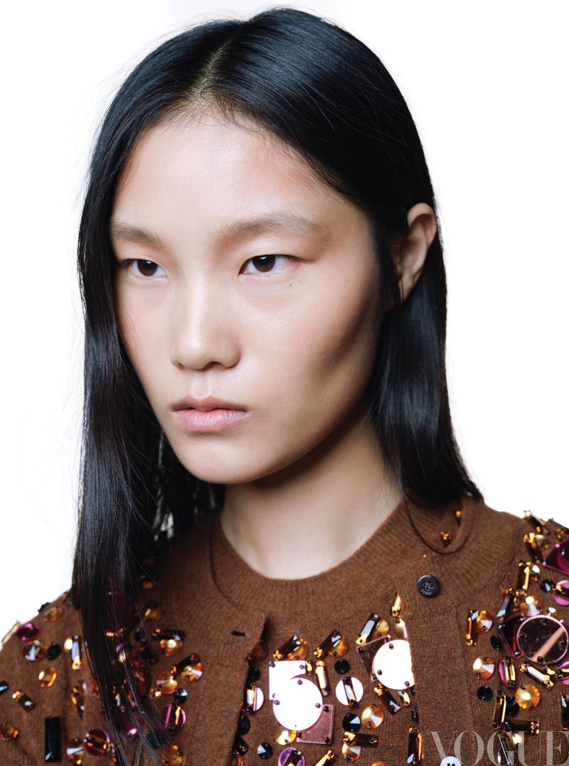 Portrait of an Asian female model with dark hair and a brown embellished cardigan, posing in a studio with a white background.